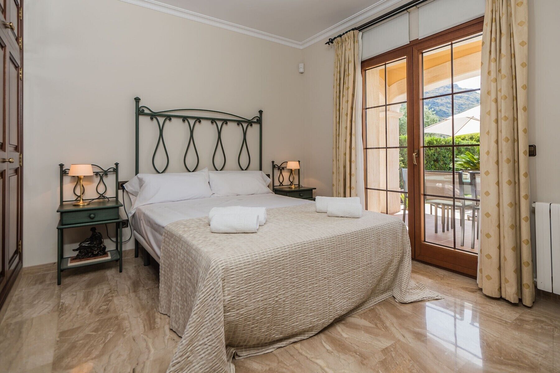 Un dormitorio con cama, dos mesitas de noche con lámparas, un ventanal con cortinas y vistas al exterior.