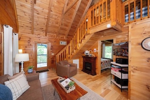 A wooden cabin interior with a sofa, armchair, fireplace, and a staircase leading to an upper level.