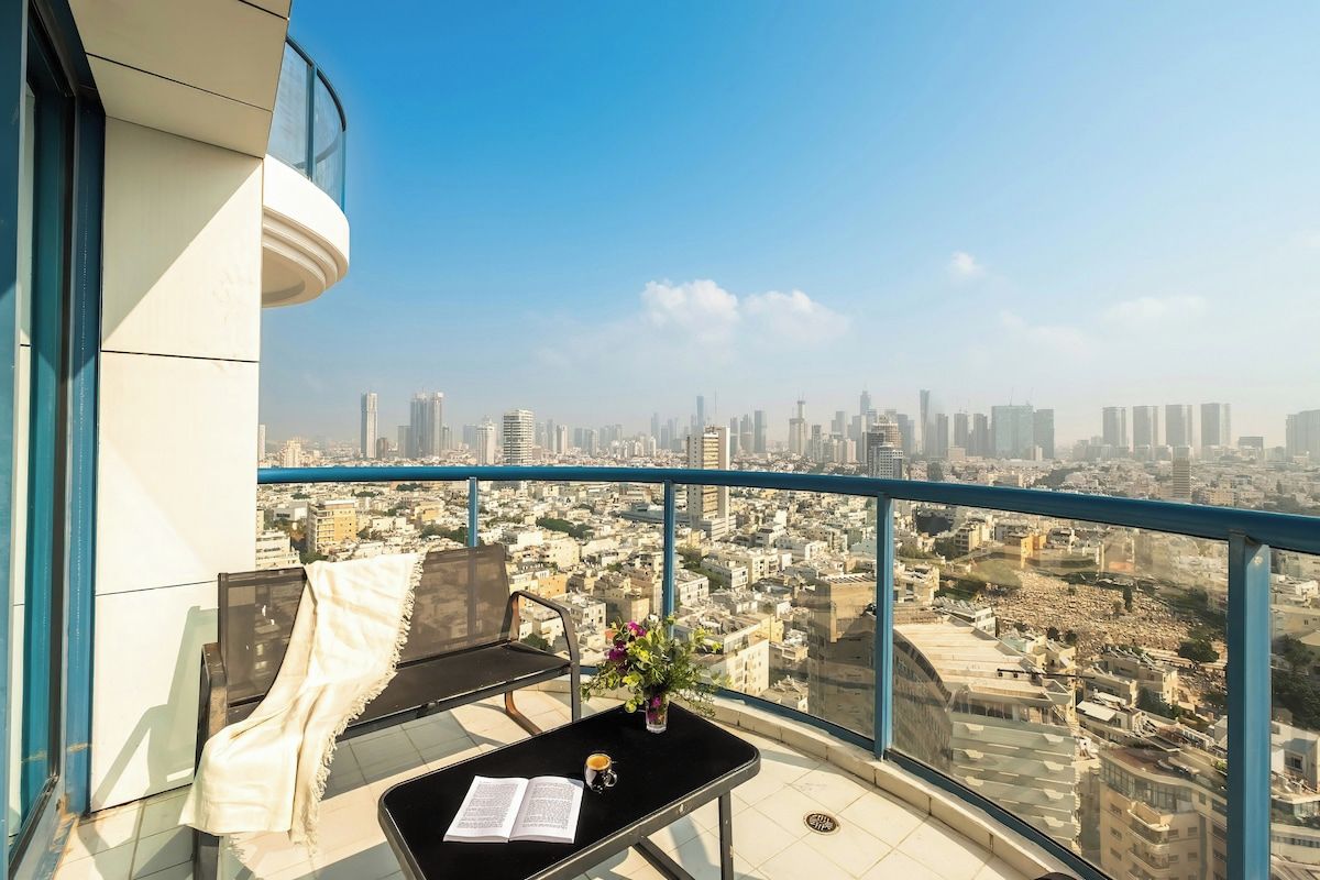Un balcon avec vue sur la ville, une table avec un livre et une tasse, et une chaise avec une couverture.