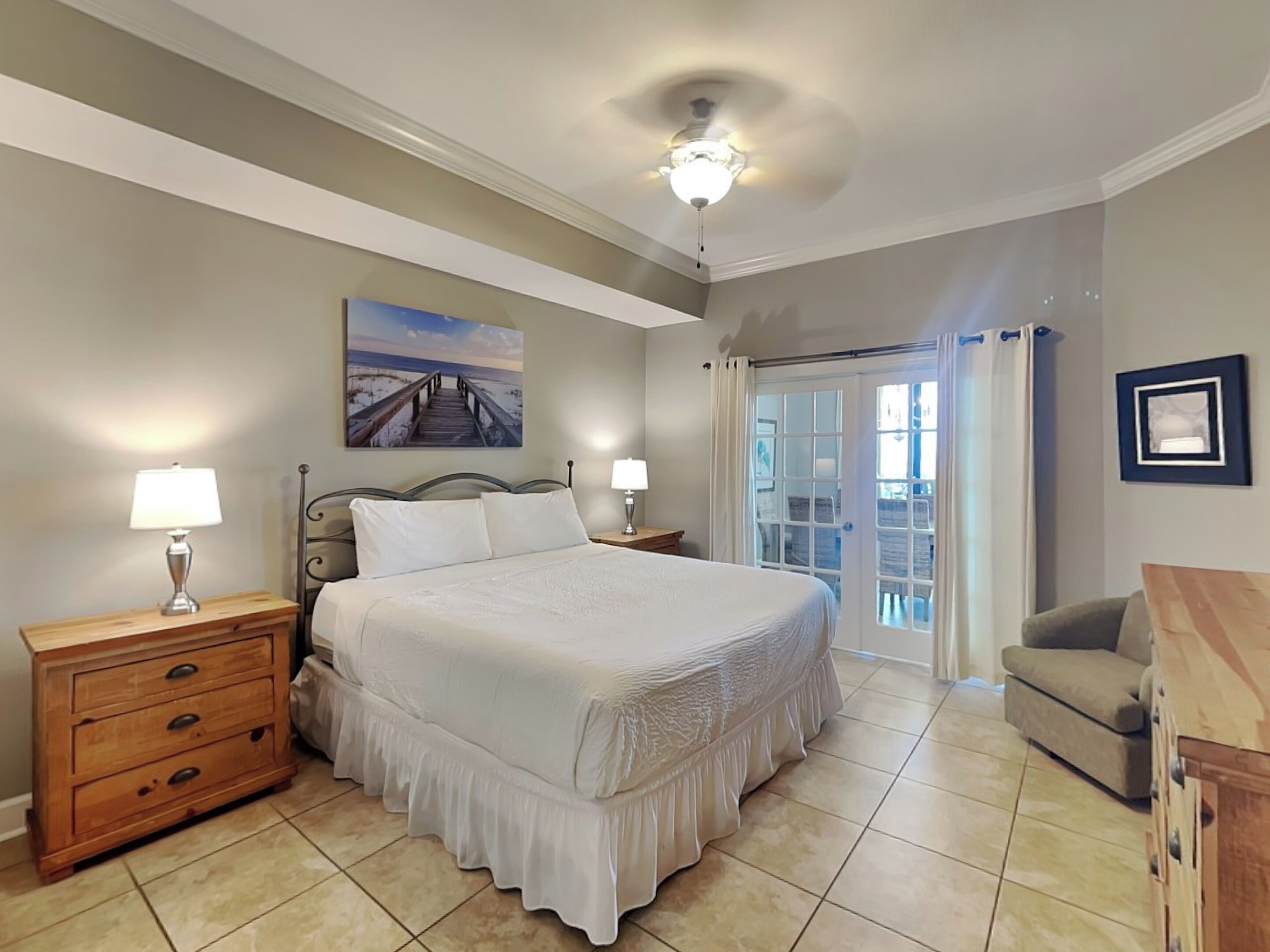 Une chambre avec un lit, deux tables de chevet avec des lampes, une commode en bois, un tableau au mur et une vue à travers une porte vitrée.