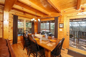 A wooden cabin interior with a dining table set for four, surrounded by chairs, and a large window offering a view of the outdoors. - Private, Beautiful Views! Pool Table, Hot Tub, Arcade Game! (Sevierville)