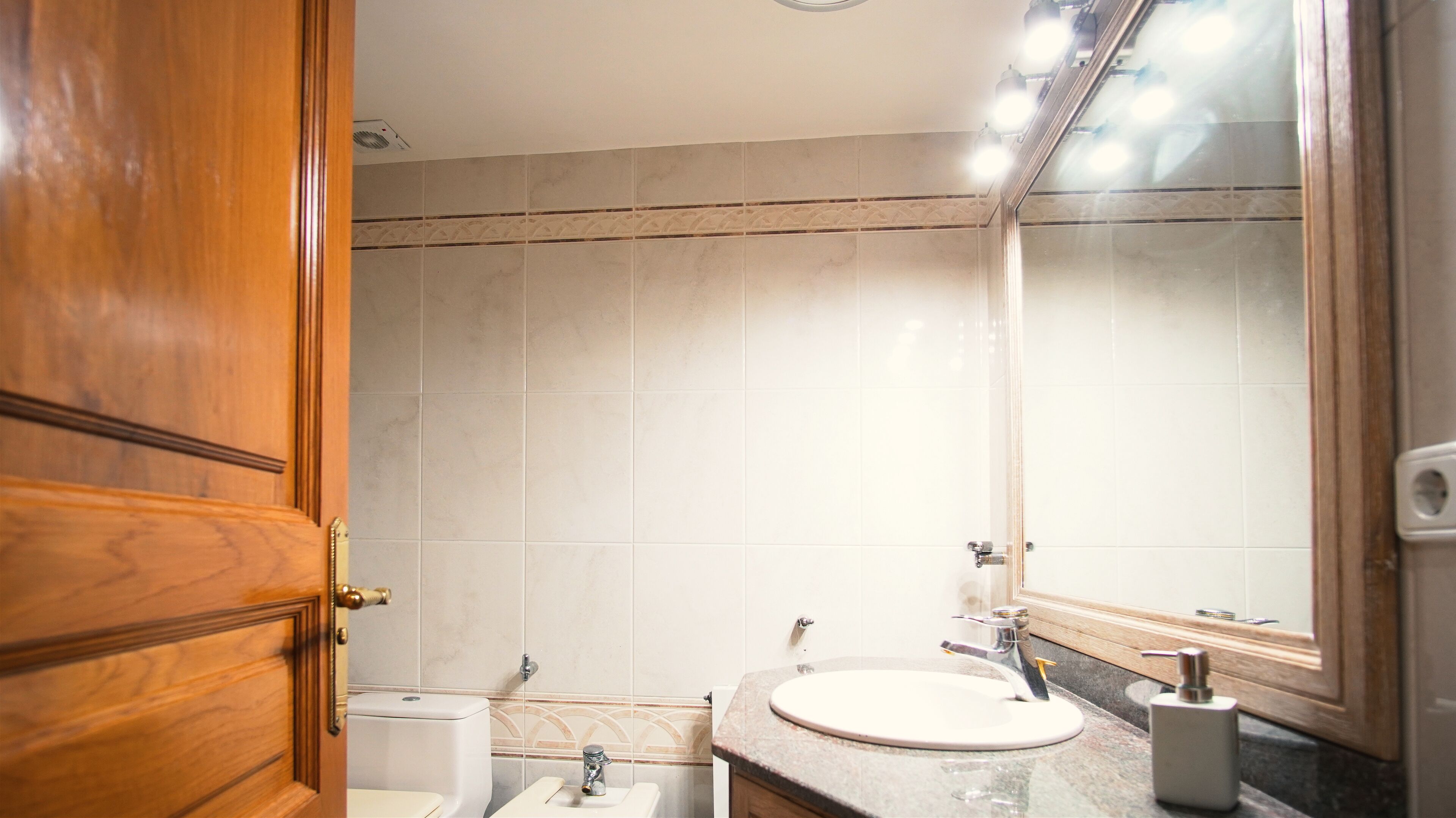 A bathroom with a wooden door, white tiles, a sink, and a mirror.