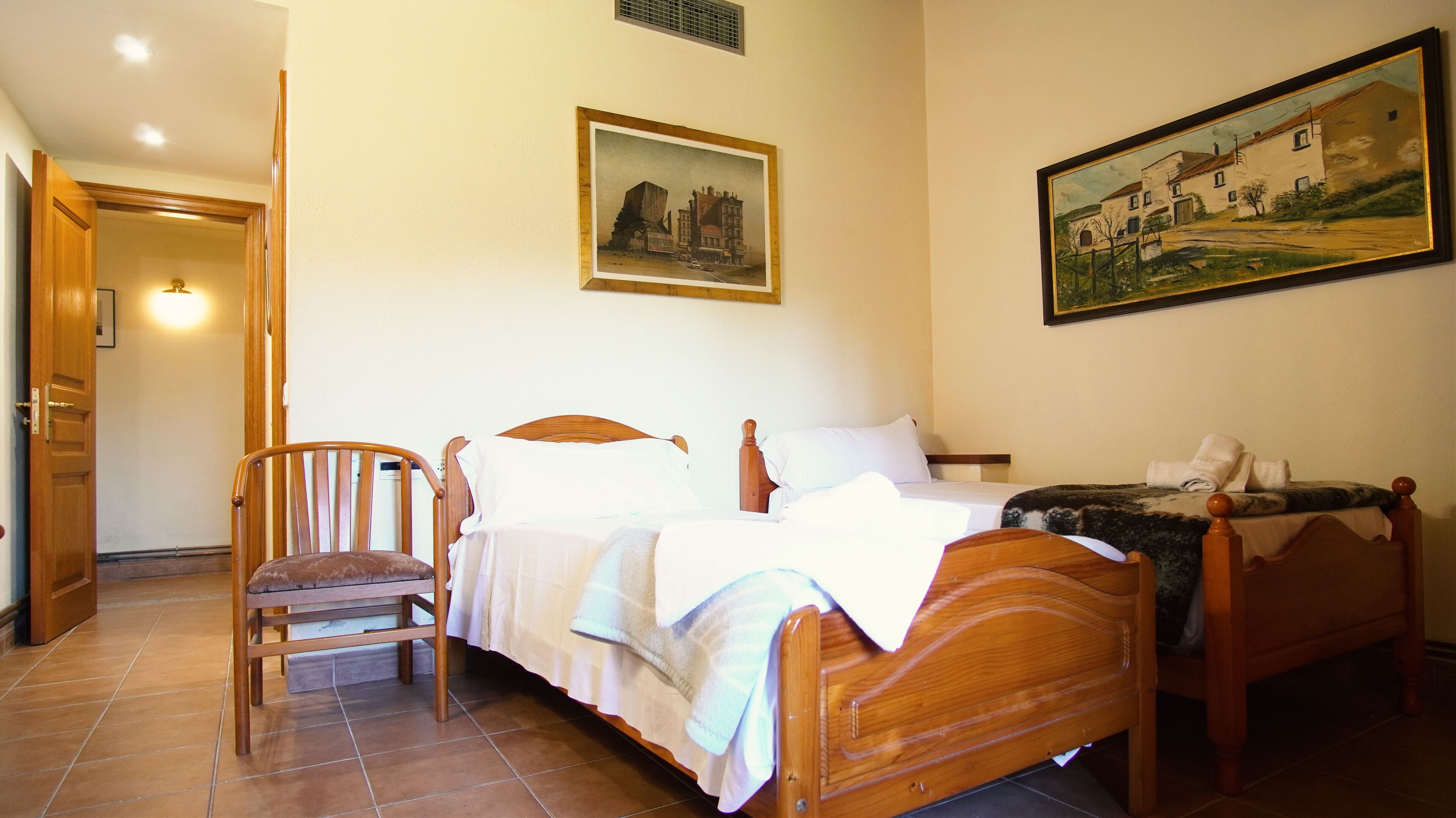 A bedroom with a wooden bed, a chair, a painting, and a door leading to another room.