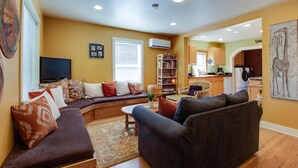 A living room with a sofa, a coffee table, and a bookshelf. There is a television on a stand and a window with blinds. - Aspen House- Sweet, Modern Downtown Cottage, 100+mbps Wifi, A/C, Walk Everywhere (Bend)