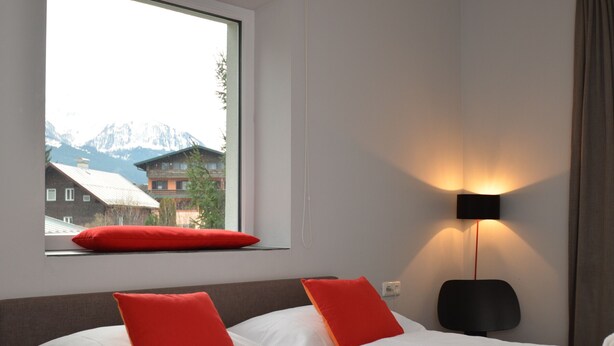 Une chambre d’hôtel avec un lit, deux oreillers rouges, une fenêtre donnant sur les montagnes et une lampe fixée au mur.