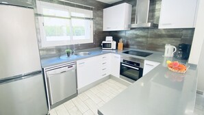 A modern kitchen with white cabinets, stainless steel appliances, a window with blinds, and a countertop with a fruit basket.