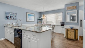 A modern kitchen with white cabinetry, a granite countertop, a dishwasher, and a dining area with a table and chairs.