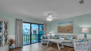 A living room with a large sliding glass door leading to a balcony, a ceiling fan, a sofa set, and a painting on the wall.