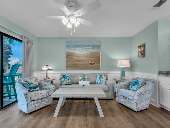 Un salon doté d’un ventilateur de plafond, d’un tableau sur le thème de la plage et de deux fauteuils ornés de motifs marins.