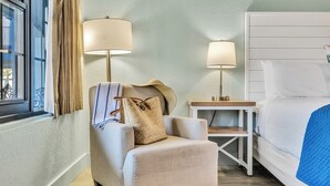 Une chambre d’hôtel avec un lit, une chaise avec une lampe, une table de chevet et une fenêtre avec des rideaux.