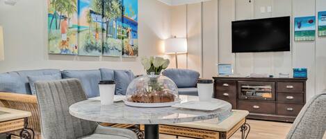 A dining area with a round table, grey upholstered chairs, a glass-top table, a ceiling fan, and a wall-mounted flat-screen TV.
