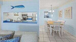 Eine moderne Küche mit weißen Schränken, einer blauen Arbeitsplatte und einem Wandbild mit Fischen. Es gibt einen Essbereich mit einem Holztisch und weißen Stühlen.