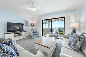 A living room with a TV, a ceiling fan, a large window, and a view of the ocean. - Beach Front 2 bed, 2 bath at Sanibel Moorings #1112 (Sanibel)