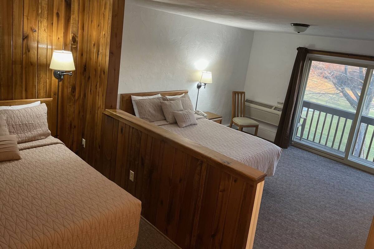 Una habitación con una cama de madera, una silla, un escritorio con lámpara y un ventanal grande.