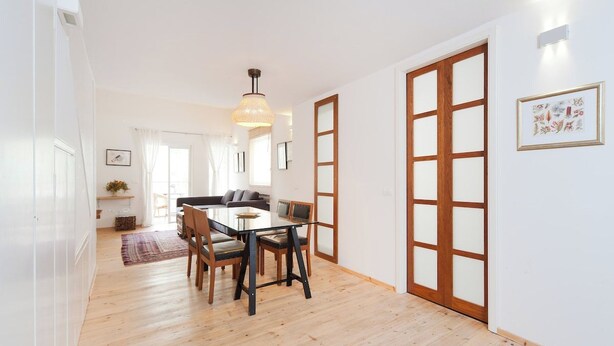 A dining area with a wooden table and chairs, a hanging light fixture, and a sliding glass door. - Stylish 1BR with Balcony — Steps to Beach & City (Tel Aviv-Yafo)