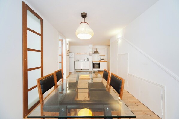 Un área de comedor con una mesa de vidrio y sillas de madera, una cocina al fondo y una lámpara colgante arriba.