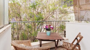 A balcony with wicker furniture, a table with a flower pot, and a view of trees and buildings. - Stylish 1BR with Balcony — Steps to Beach & City (Tel Aviv-Yafo)
