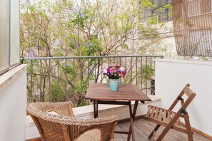 Un balcone con mobili in vimini, un tavolo con un vaso di fiori e vista su alberi e edifici.