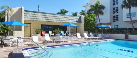 A swimming pool with lounge chairs and umbrellas, a building with a green roof, and palm trees.