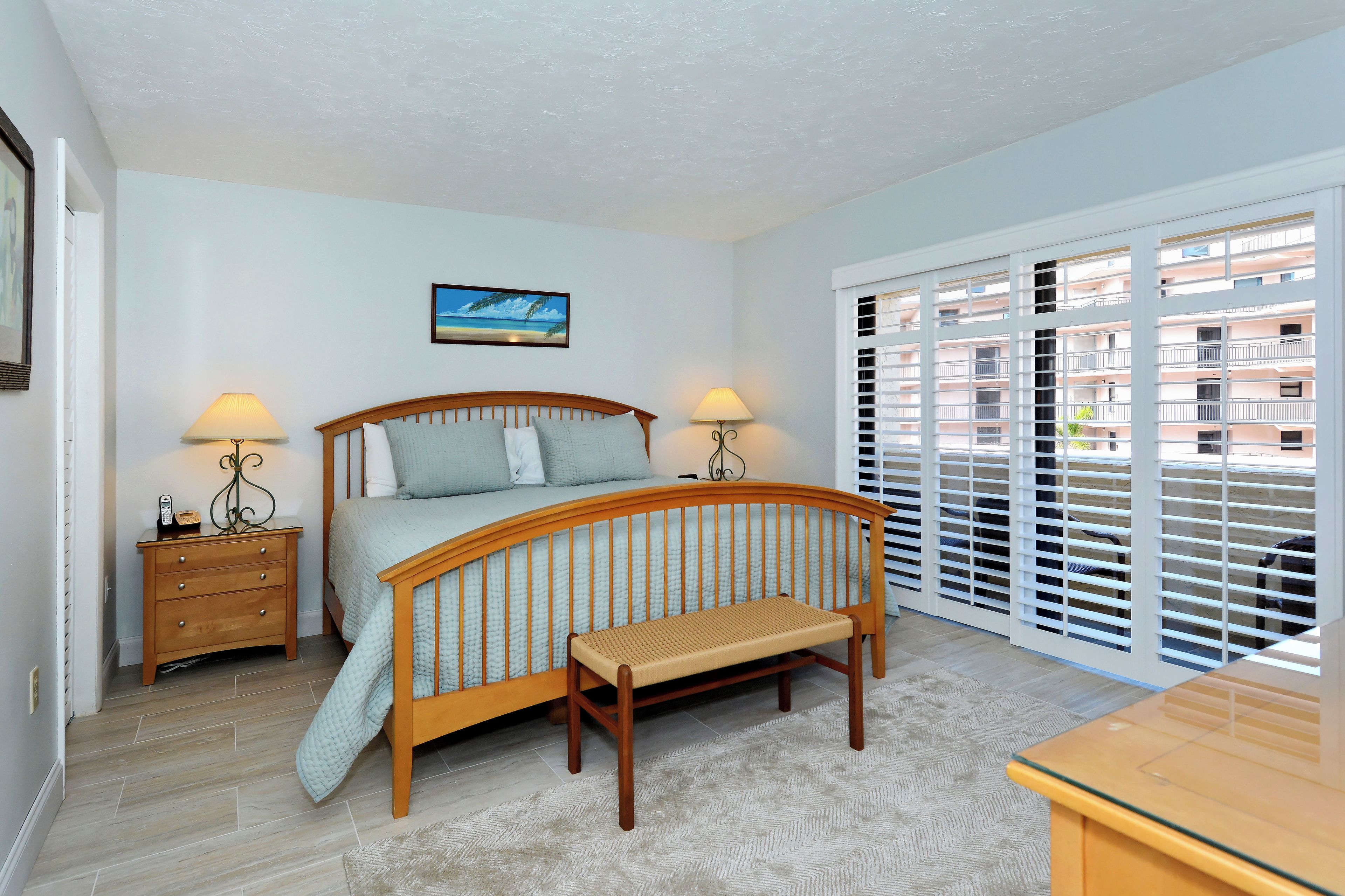 A bedroom with a wooden bed, two bedside tables with lamps, a bench, a large window with blinds, and a painting on the wall.