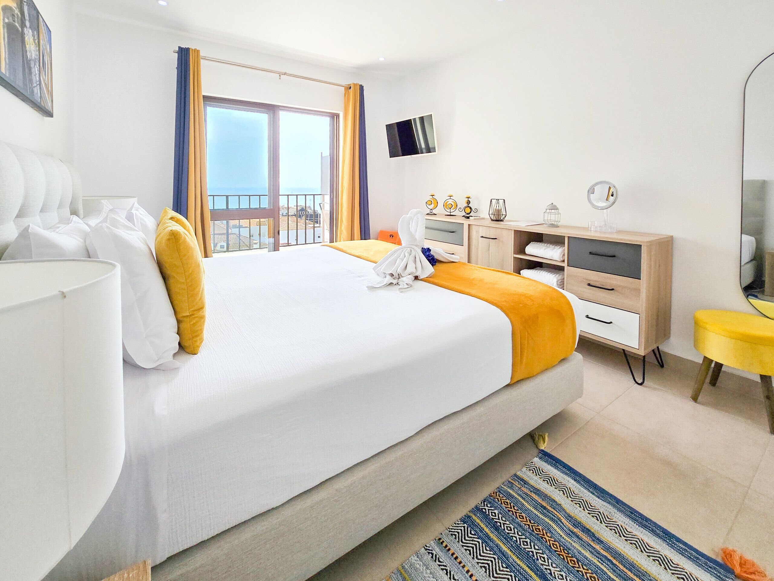 A hotel room with a large bed, a desk with a chair, a small table with a mirror, and a view of the sea through the window.