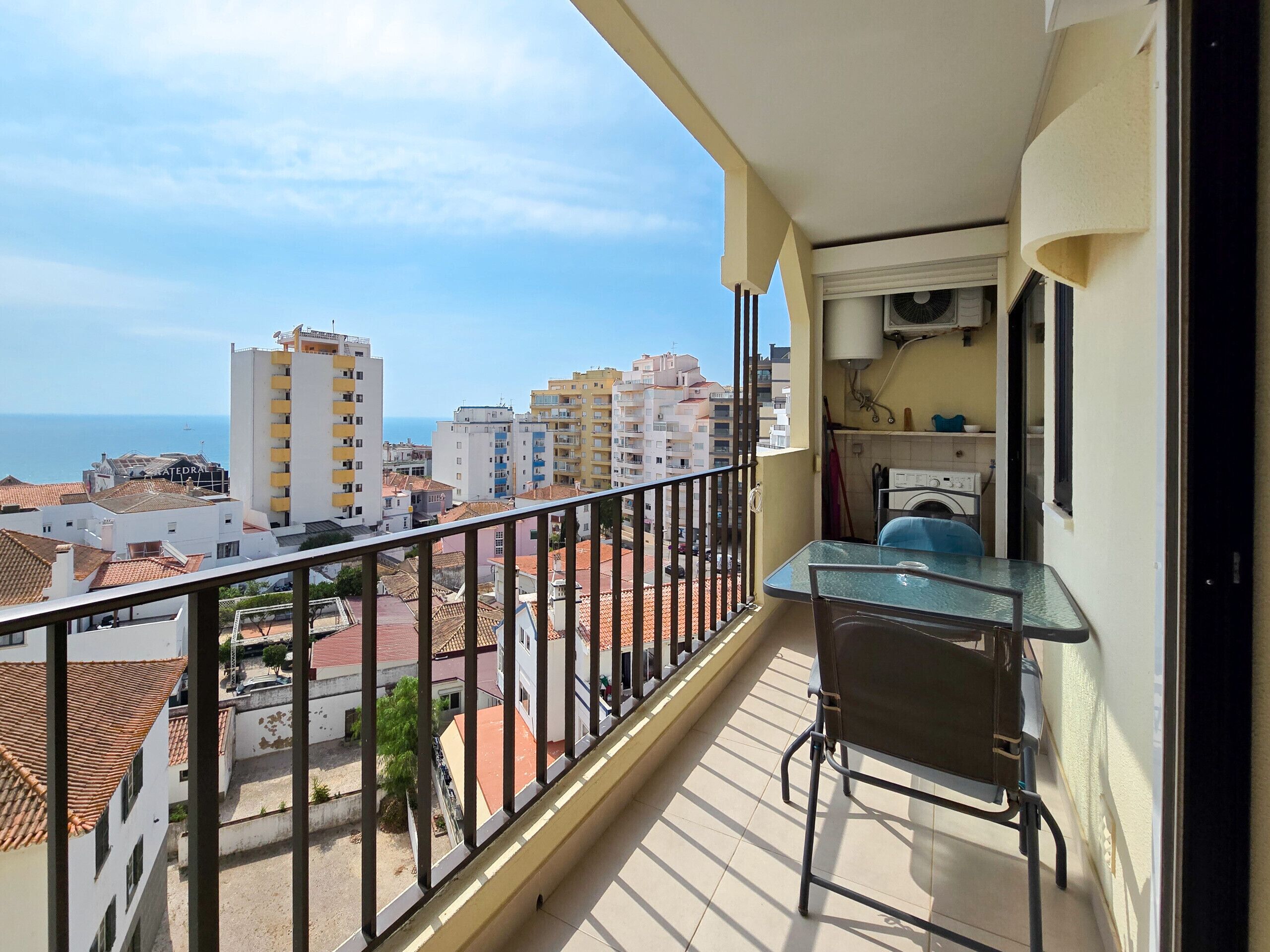 A balcony with a table and chair, overlooking a coastal cityscape with buildings and the sea.