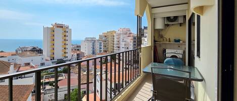 A balcony with a table and chair, overlooking a coastal cityscape with buildings and the sea.