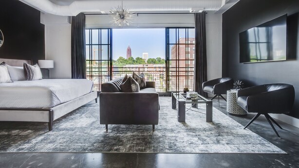 A modern hotel room with a large bed, a sofa, a coffee table, and a TV. The room has large windows with a view of buildings and trees. - Ponce City Market Studio · Heart of Atlanta · BeltLine (Atlanta)