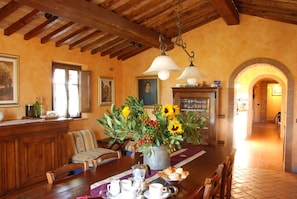Comedor con mesa de madera, sillas y un florero de girasoles. Se observa una chimenea y un banco de madera al fondo.