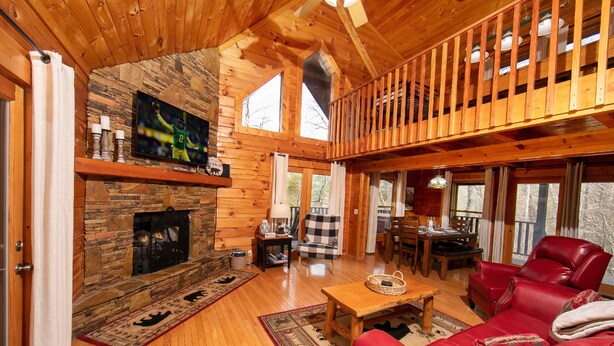 A spacious living room with a stone fireplace, wooden floors, a red leather sofa, and a balcony with a wooden railing. - Private Cabin, Hot Tub, Pool Table, Walking Distance to Fishing Pond (Sevierville)