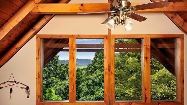 Una habitación con techo de madera y ventilador de techo. Hay tres ventanas amplias con vista a un bosque y montañas.