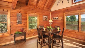 Interior de una cabaña de madera con una mesa de comedor, sillas y una ventana que da a un bosque.