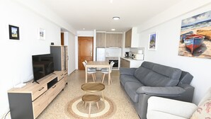 Uma sala de estar moderna com um sofá cinzento, um móvel de TV de madeira, uma mesa de jantar com cadeiras e uma área de cozinha ao fundo.
