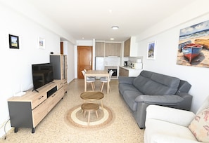 Een moderne woonkamer met een grijze bank, een houten tv-meubel, een eettafel met stoelen en een keuken op de achtergrond.