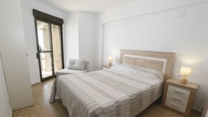 Quarto com cama, mesas de cabeceira, uma cadeira e porta de vidro deslizante que dá para área externa.