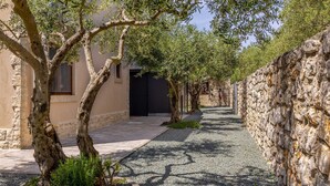 Une ruelle étroite bordée d’oliviers et de murs de pierre, qui mène à un bâtiment doté d’une porte sombre.