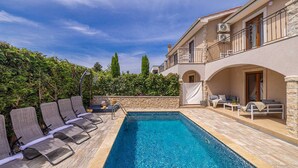 Un espace aménagé au bord de la piscine, avec des chaises longues, un mur en pierre et un bâtiment à deux étages doté de balcons.