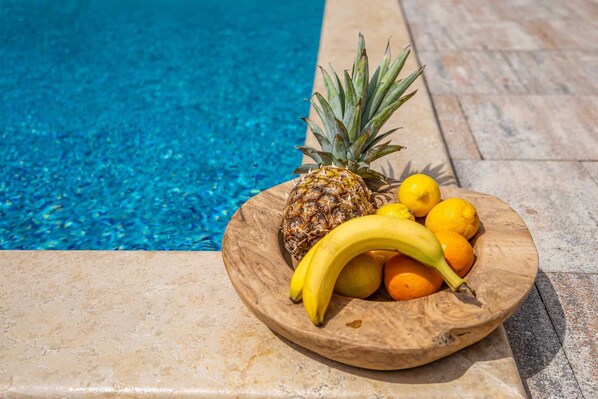 Een houten schaal met ananas, bananen en sinaasappels bij een zwembad.