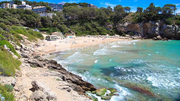 A coastal beach with rocky terrain, clear turquoise waters, and a backdrop of buildings and greenery. - Apartment with Pool in Cala Crancs area · UHC CASAS BLANCAS 121 (Salou)
