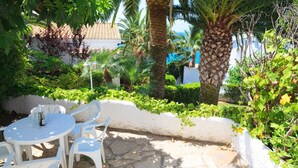 An outdoor patio with a round table and white plastic chairs, surrounded by lush greenery and palm trees. - Apartment with Pool in Cala Crancs area · UHC CASAS BLANCAS 121 (Salou)