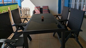 En overdækket terrasse med et bord og stole, en blå væg og udsigt til stranden.