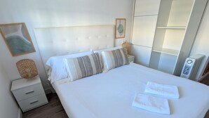En pænt redt seng med hvide sengetæpper og stribede puder, et natbord med lampe, og en stor garderobe.