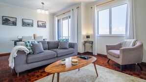 A modern living room with a grey sofa, a wooden coffee table, and a beige armchair. There are large windows with white curtains, a small side table, and wall-mounted artwork. - Sea View - By WeHostMadeira (Câmara de Lobos)