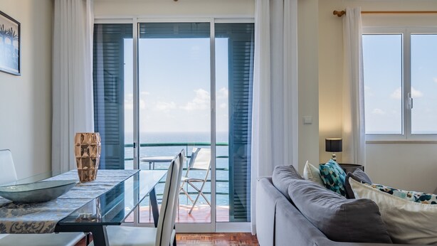 A modern living room with a sofa, a dining table with chairs, and a view of the sea through large glass doors. - Sea View - By WeHostMadeira (Câmara de Lobos)