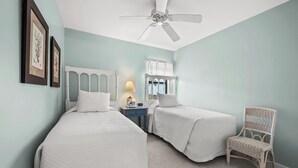 Une chambre avec deux lits, un ventilateur de plafond, une table de chevet avec une lampe et un fauteuil en osier.
