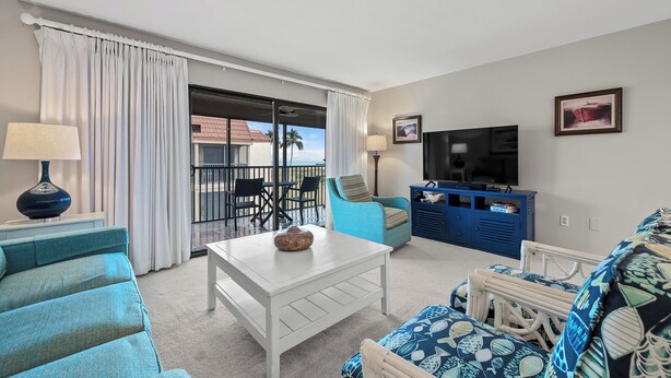 Un salon avec une table basse blanche, un canapé bleu, un fauteuil bleu, un téléviseur et un balcon offrant une vue sur l’océan.
