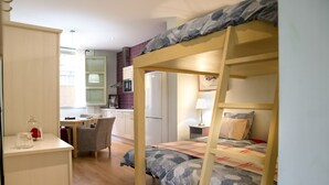 Ein Etagenbett mit Tarnmuster-Bettwäsche, ein Schreibtisch mit Stuhl und eine Küchenzeile im Hintergrund.