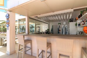 An outdoor bar area with wicker bar stools, a counter, hanging wine glasses, and shelves with bottles and plants.