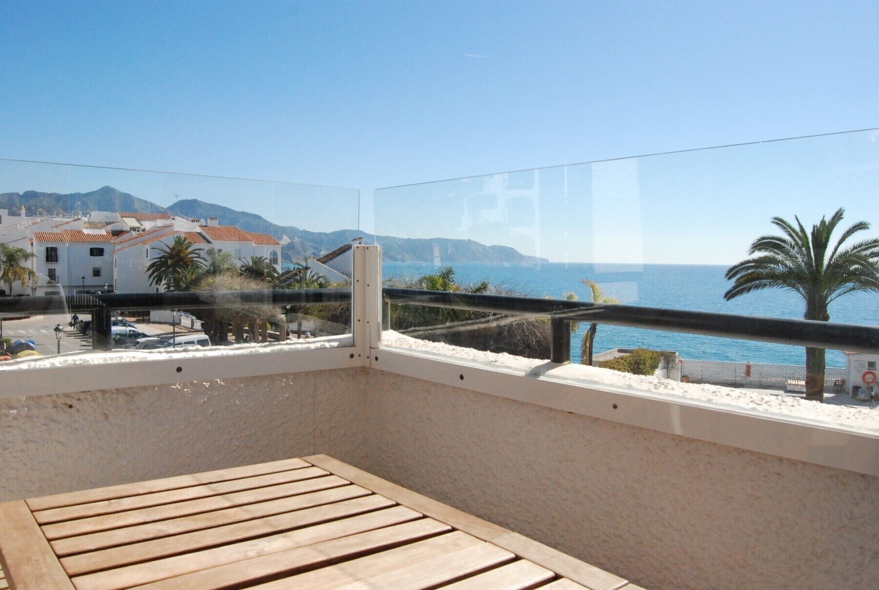 A balcony with a wooden table, a glass railing, and a view of the sea and buildings.