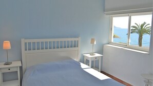 A bedroom with a bed, a nightstand, a lamp, a window with a view of the sea, and a small table.